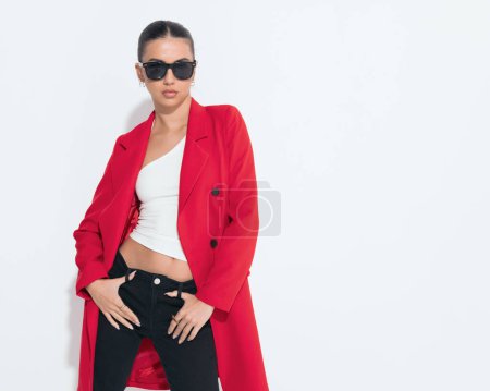 Photo for Sexy young girl with sunglasses in red coat holding black pants and posing in a cool way on white background - Royalty Free Image