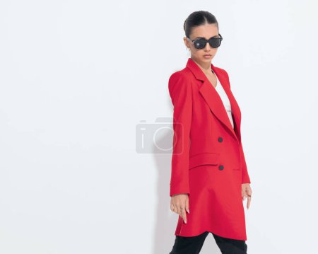 Téléchargez les photos : Femme de mode sexy avec des lunettes de soleil en manteau rouge regardant vers l'avant et posant tout en marchant devant fond blanc - en image libre de droit