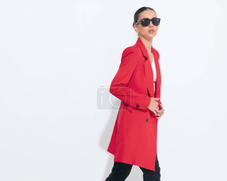 Téléchargez les photos : Vue latérale de la femme de mode cool avec des lunettes de soleil boutonnage manteau rouge tout en marchant et en regardant loin sur fond blanc - en image libre de droit