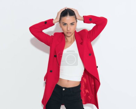 Téléchargez les photos : Cool fille de mode en manteau rouge ajustant la coiffure de chignon et regardant vers l'avant tout en posant sur fond blanc - en image libre de droit