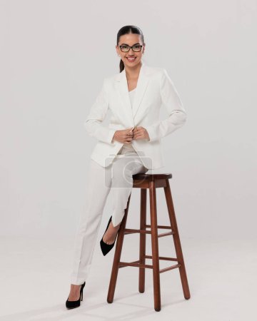 Photo for Happy elegant woman with glasses adjusting white suit and smiling while sitting on wooden chair in front of grey background - Royalty Free Image
