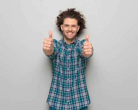 Téléchargez les photos : Homme décontracté enthousiaste avec des lunettes souriant et faisant les pouces geste devant fond gris - en image libre de droit