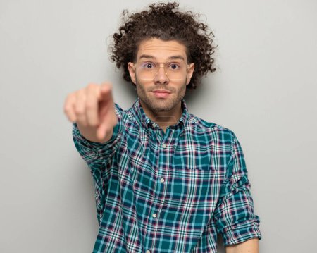 Foto de Hombre guapo con el pelo rizado mirando hacia adelante y señalando el dedo hacia adelante elegir mientras está de pie delante de fondo gris - Imagen libre de derechos