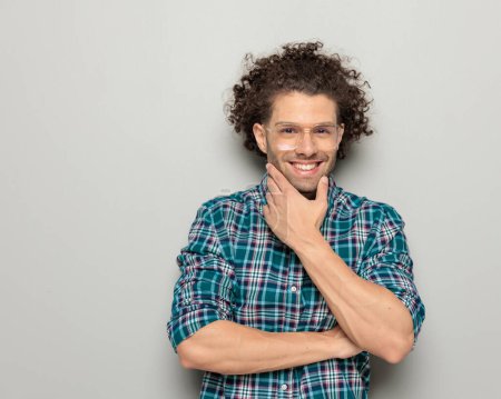 Photo for Portrait of happy young man with curly hair wearing checkered shirt and glasses, folding arms, touching chin and dreaming in front of grey background - Royalty Free Image
