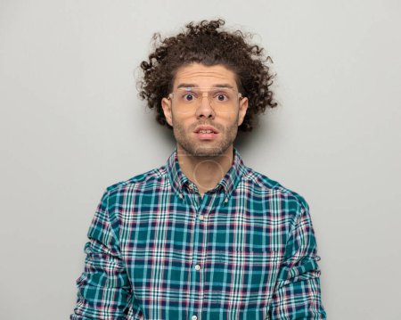 Photo for Amazed young guy with glasses in checkered shirt looking forward and being surprised in front of grey background - Royalty Free Image