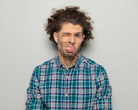 Photo for Portrait of funny casual guy with curly hair and glasses wearing checkered shirt making faces, sticking out tongue and posing in a fun way on grey background - Royalty Free Image