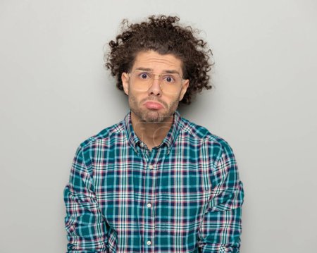 Photo for Portrait of casual curly hair man with glasses making a cute upset face and looking forward, begging in front of grey background - Royalty Free Image