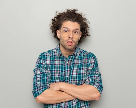 Téléchargez les photos : Portrait de jeune homme surpris avec des lunettes en chemise à carreaux croisant les bras et faisant un visage stupide devant fond gris - en image libre de droit