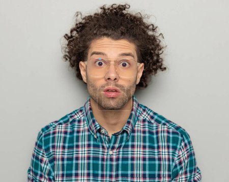 Photo for Shocked young man with glasses making a surprised face with big eyes and looking forward in front of grey background - Royalty Free Image