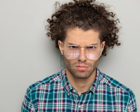 Téléchargez les photos : Attrayant jeune homme avec des lunettes en chemise à carreaux regardant vers l'avant et faire un joli visage bouleversé devant fond gris - en image libre de droit