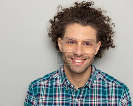 Foto de Imagen de cerca de un joven feliz con el pelo rizado con gafas y camisa a cuadros mirando hacia adelante y sonriendo sobre fondo gris - Imagen libre de derechos