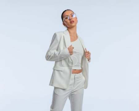 Foto de Sexy mujer de negocios elegante en traje blanco con gafas de ajuste traje mientras posa de una manera segura delante de fondo gris - Imagen libre de derechos