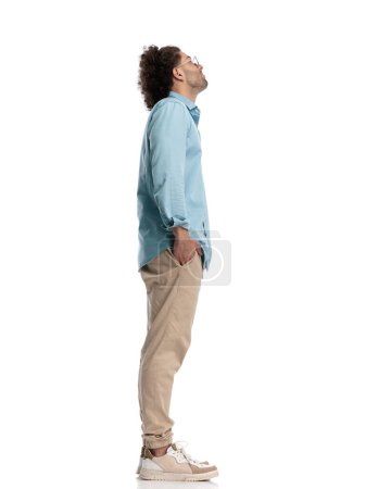 Photo for Young casual man waiting in line and looking up with hands in pockets on isolated background - Royalty Free Image