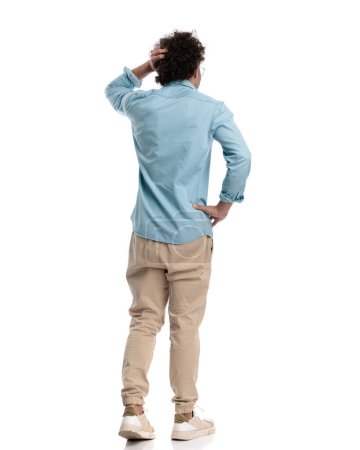 Photo for Rear view of confused young man holding his head and hips while standing on white background - Royalty Free Image