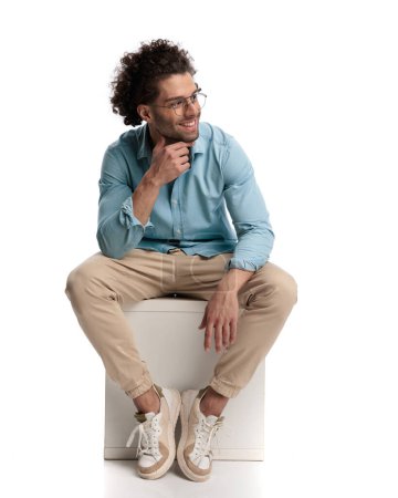 Photo for Pensive casual man sitting on a crate and looking to side on white background - Royalty Free Image