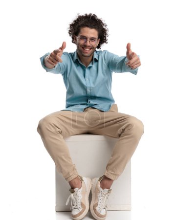 Photo for Joyful casual man sitting on a chair on white background and making thumbs up sign - Royalty Free Image