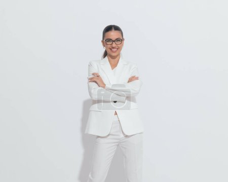 Téléchargez les photos : Femme d'affaires heureuse avec des lunettes en costume blanc bras croisés et riant tout en posant devant fond gris - en image libre de droit
