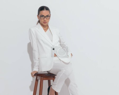 Téléchargez les photos : Femme d'affaires confiante avec des lunettes en costume blanc tenant la main dans la poche et regardant vers l'avant devant fond gris - en image libre de droit