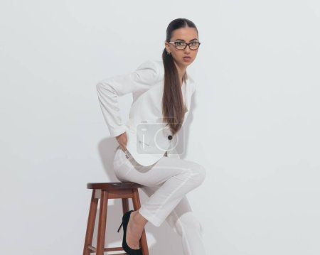 Foto de Portait de mujer de moda elegante con gafas cogidas de la mano en el bolsillo y sentado en silla de madera en frente de fondo gris - Imagen libre de derechos