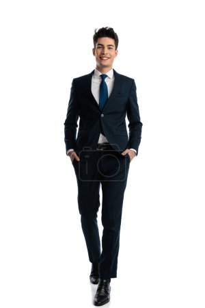 Photo for Full length picture of happy elegant man in navy blue suit stepping with hands in pockets and smiling in front of white background - Royalty Free Image