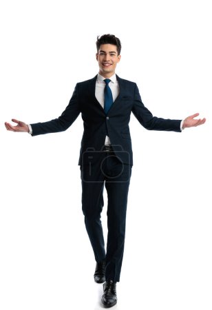 Foto de Atractivo joven empresario en traje azul marino caminando y presentando con los brazos abiertos en frente de fondo blanco, cuerpo completo - Imagen libre de derechos