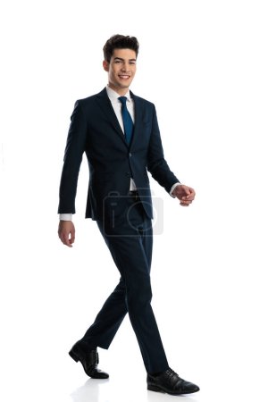 Photo for Full body picture of happy elegant businessman in navy blue suit smiling and walking in front of white background - Royalty Free Image