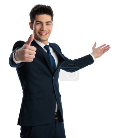 Foto de Feliz joven empresario haciendo pulgares hacia arriba signo, sonriendo y mostrando a un lado en frente de fondo blanco - Imagen libre de derechos