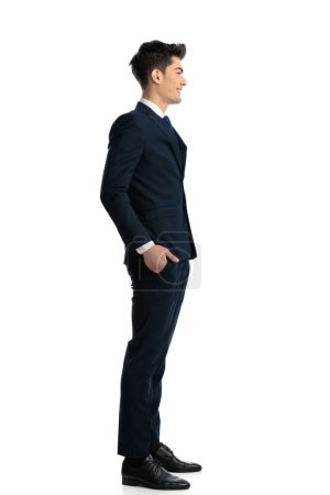 Foto de Foto vista lateral de hombre elegante guapo en traje azul marino con las manos en bolsillos esperando en línea y sonriendo delante de fondo blanco - Imagen libre de derechos