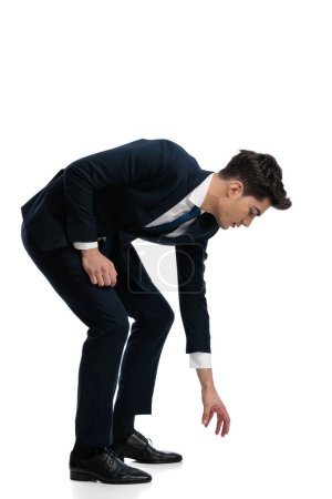 Foto de Vista lateral del elegante hombre de la moda inclinándose y agarrando algo del suelo frente al fondo blanco - Imagen libre de derechos