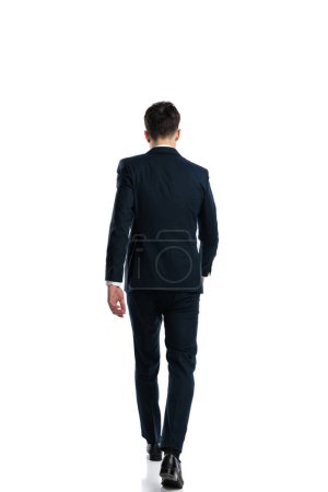 Téléchargez les photos : Vue arrière photo de jeune homme élégant en costume bleu marine marchant devant fond blanc - en image libre de droit