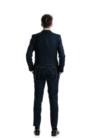Photo for Rear view of elegant man in navy blue suit holding hands in pockets and standing in front of white background - Royalty Free Image
