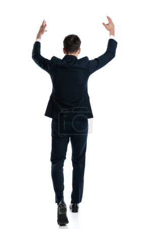 Foto de Vista trasera de empresario elegante emocionado con los brazos por encima de la cabeza caminando y animando la victoria en frente de fondo blanco - Imagen libre de derechos