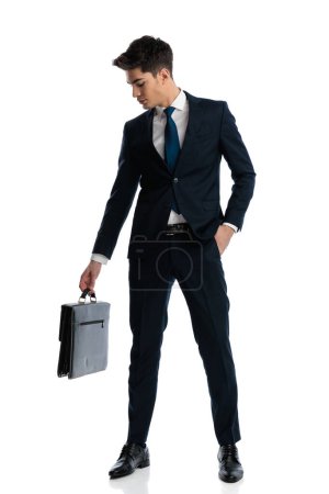 Foto de Imagen de cuerpo completo de atractivo hombre de negocios elegante en traje azul marino mirando hacia abajo a su maleta mientras está de pie con la mano en el bolsillo sobre fondo blanco - Imagen libre de derechos