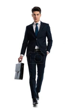 Foto de Imagen de cuerpo completo de hombre de negocios sexy confiado sosteniendo la maleta y caminando con las manos en los bolsillos delante del fondo blanco - Imagen libre de derechos