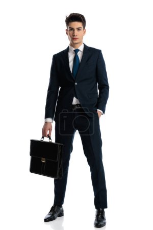 Foto de Imagen de cuerpo completo de atractivo joven hombre de negocios en traje azul marino bolsa de sujeción y posando con la mano en el bolsillo delante del fondo blanco - Imagen libre de derechos