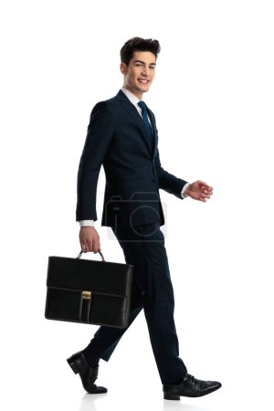 Téléchargez les photos : Vue latérale d'élégant jeune homme d'affaires en costume bleu marine avec valise marchant et souriant devant fond blanc - en image libre de droit