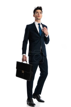 Photo for Full body picture of handsome young man looking up while adjusting suit and holding suitcase, walking confidently in front of white background - Royalty Free Image