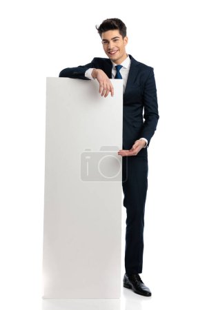 Photo for Proud businessman in navy blue suit showing and presentiny white empty board, smiling in front of white background - Royalty Free Image