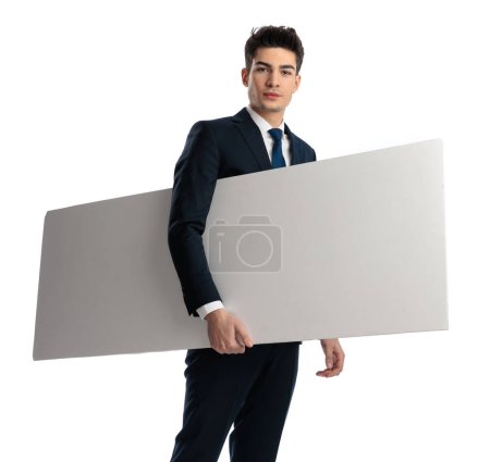 Téléchargez les photos : Homme sexy confiant en costume tenant tableau blanc vide sous son bras et regardant vers l'avant en face de fond blanc - en image libre de droit
