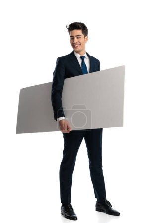 Téléchargez les photos : Homme d'affaires excité avec signe vide sous son bras regardant de côté et riant tout en se tenant devant fond blanc - en image libre de droit