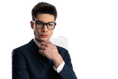 Téléchargez les photos : Portrait de jeune homme réfléchi avec des lunettes en costume élégant touchant menton et la pensée en face de fond blanc - en image libre de droit