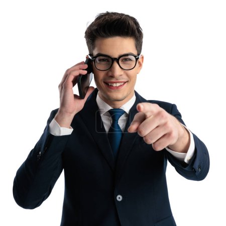 Téléchargez les photos : Heureux homme d'affaires élégant avec des lunettes ayant une conversation téléphonique, souriant et pointant le doigt vers l'avant devant fond blanc - en image libre de droit