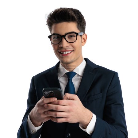 Téléchargez les photos : Portrait d'homme d'affaires heureux en costume tenant mobile et écrivant un message tout en souriant devant fond blanc - en image libre de droit