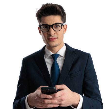 Photo for Portrait of attractive businessman with glasses holding phone and writing a message while squinting in front of white background - Royalty Free Image