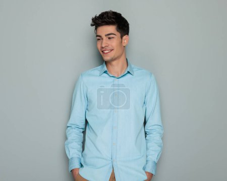 hombre casual feliz en camisa de mezclilla azul mirando hacia otro lado y riendo mientras toma de la mano en bolsillos delante de fondo blanco