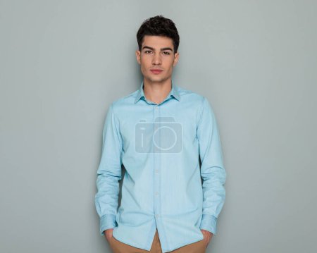 Photo for Portrait of attractive young man in blue denim shirt holding hands in pockets and looking forward in front of grey background - Royalty Free Image
