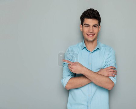 Photo for Cute casual man in denim shirt crossing arms, pointing finger to side while smiling in front of white background - Royalty Free Image