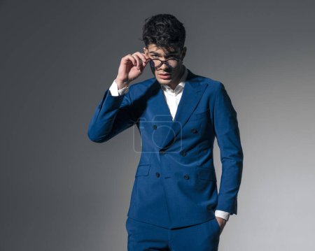 Téléchargez les photos : Homme de mode sexy et détendu fixant ses lunettes tout en se tenant la main dans la poche sur fond sombre - en image libre de droit