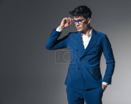 Photo for Confident businessman with hands in pockets arranging glasses and looking down to side while standing on dark background - Royalty Free Image