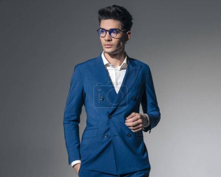 Foto de Sexy hombre de negocios vistiendo doble pecho azul traje y gafas es la celebración de bolsillos, mientras que mirando a un lado sobre fondo gris - Imagen libre de derechos